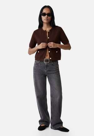 Mujer con chaqueta marrón de manga corta con botones, vaqueros anchos de color gris oscuro, cinturón negro, zapatos negros y gafas de sol negras sobre fondo blanco.