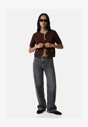 Kvinde iført brun kortærmet jakke med knapper, mørkegrå vidde jeans, sort bælte, sorte sko og sorte solbriller mod hvid baggrund.