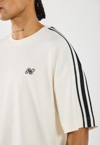 Chemise crème à manches courtes avec des accents rayés noir et blanc sur les épaules et un petit logo noir brodé sur la poitrine.
