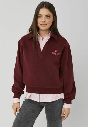 Sweatshirt bordeaux avec col et fermeture éclair mi-haute, arborant un logo rose brodé. Porté par-dessus une chemise rayée rose clair et un jean gris.