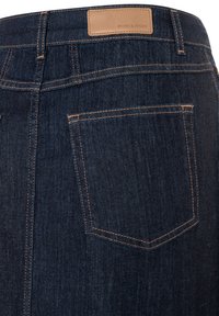 Jean en denim bleu foncé avec une étiquette en cuir camel à la taille et des coutures visibles. Une seule poche arrière est visible, avec un design pointu.