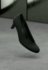 Zwarte leren pump met een ronde neus en een lage, stevige hak; gladde textuur en minimalistisch ontwerp, zwevend boven een grijze oppervlakte.