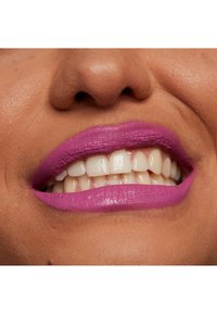 Primo piano di labbra con rossetto viola brillante opaco, in contrasto con denti chiari e pelle texturizzata. Focus sul colore e sull'applicazione liscia.