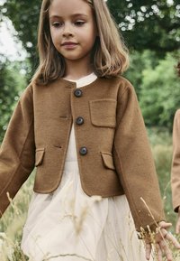 Hnědá vlněná bunda s kulatým výstřihem, čtyřmi černými knoflíky a náprsní kapsou, kombinovaná s bílými šaty s průhlednou vrstvou.