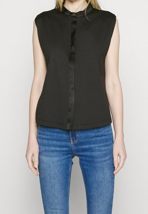 Mouwloze zwarte blouse met een mandarijnkraag en knoopsluiting aan de voorkant, gedragen met blauwe spijkerbroek door een staand persoon.