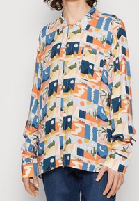 Chemise colorée à boutons avec un design patchwork comprenant des motifs abstraits, des montagnes, des cactus et des formes géométriques sur un fond clair.