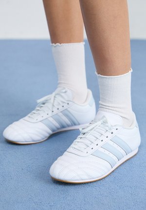 Stopy w białych butach sportowych z jasnoniebieskimi paskami i białymi prążkowanymi skarpetami na jasnoniebieskiej teksturowanej podłodze.