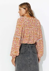 Femme avec de longs cheveux blonds portant des boucles d'oreilles en or, un blouse à manches longues à fleurs roses et une jupe en denim grise, de profil sur un fond blanc.