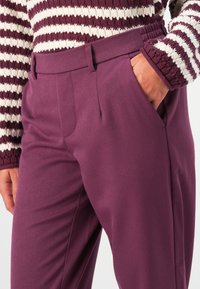 Woman wearing plum-colored tailored trousers with side pockets and a striped knit sweater, hand resting in pocket.