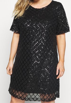 Femme portant une robe noire à manches courtes avec un motif en diamants en sequins et un col rond, se tenant devant un fond uni.