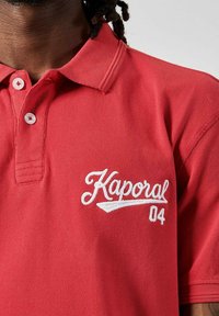 Rode polo shirt van een getextureerde stof, met een witte geborduurde "Kaporal 04" logo op de linkerkant van de borst en een standaard met knopen sluitende kraag.