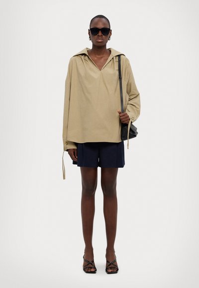 Camicia beige oversize con scollatura a V profonda e maniche larghe. Abbinata a pantaloni corti blu navy, occhiali da sole neri e una piccola borsa nera.