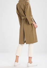 Khaki trenčkot s páskem v oblasti pasu, dlouhými rukávy a zadním rozparkem, kombinovaný s bílými zkrácenými kalhotami a bílými teniskami. Čisté linie a hladký povrch.