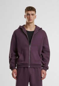 Lilla zip-up hettegenser laget av mykt stoff, med en frontal kengurulomme, snorhette og ribbede ermer og kant. Brukt med matchende bukser.