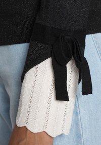 Pull en maille noir avec manches blanches texturées bordées de bords festonnés et doté d'un accent noir noué. Les matériaux incluent un fil doux et brillant.
