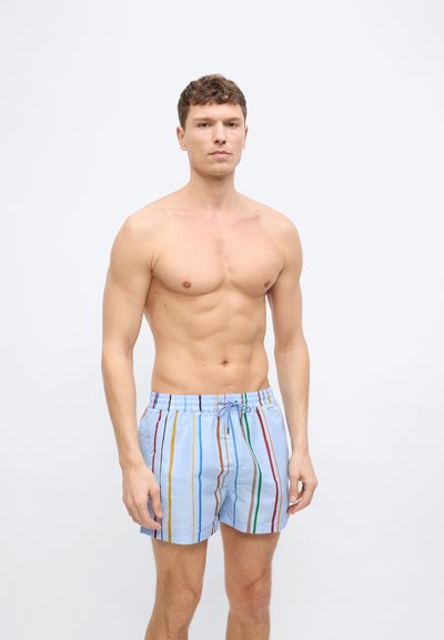 Ανδρας χωρίς πουκάμισο με κοντά σγουρά μαλλιά, φορούσε ανοιχτό μπλε μαγιό με κάθετες πολύχρωμες ρίγες, στέκεται μπροστά σε απλό λευκό φόντο.