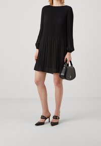 Robe noire plissée avec des manches longues et un col rond, associée à des mules noires à talons avec des accents circulaires dorés, et un petit sac à main noir.