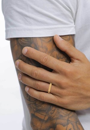 Bague en or avec une surface plate et texturée, portée sur une main reposant sur un bras tatoué. Bras exposé, portant une chemise blanche à manches courtes.