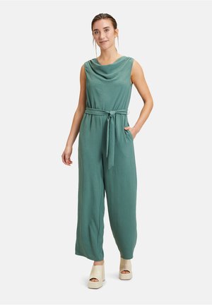 Grüner Jumpsuit mit Schalkragen und gebundenem Bund, ausgestattet mit weiten Beinen und seitlichen Taschen. Kombiniert mit cremefarbenen Plateauschuhen. Glattes, leichtes Material.