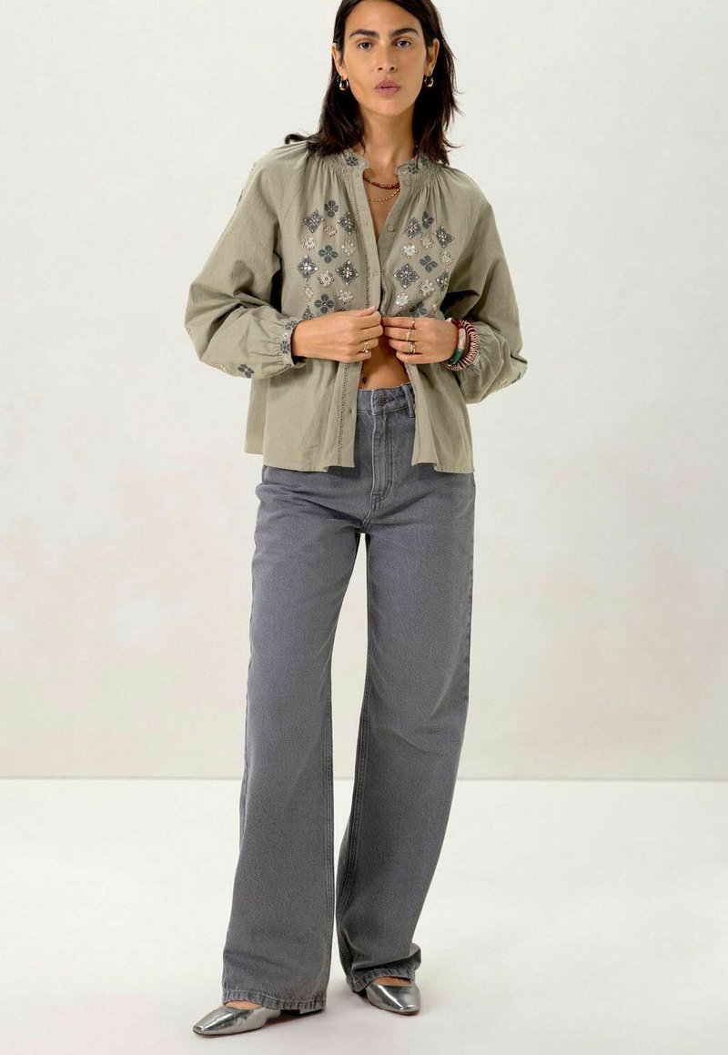 Olijfgroene button-up shirt met bloemenborduursel, gecombineerd met hoge grijze wijde jeans en zilveren puntige schoenen.