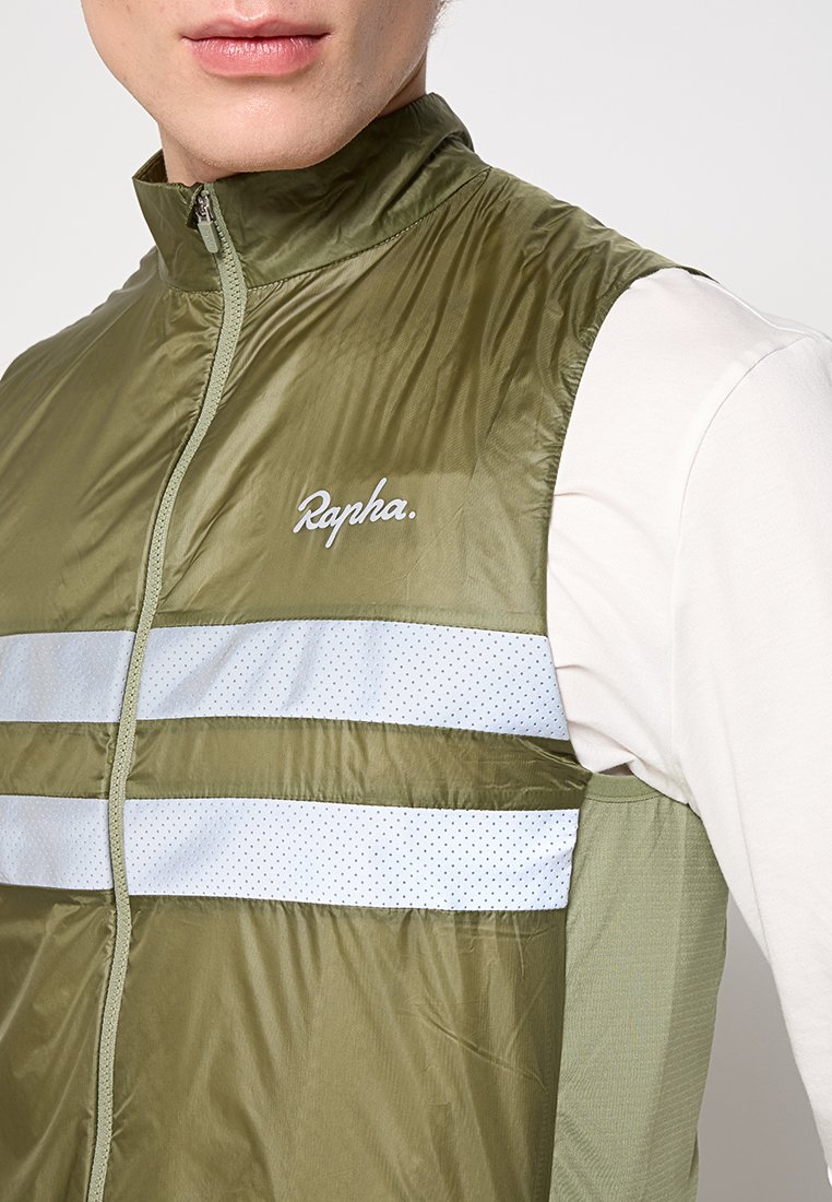 Mann trägt eine olivgrüne ärmellose Weste mit weißen reflektierenden Streifen und dem weißen "Rapha"-Logo über einem weißen Langarmshirt.
