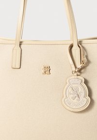 Bolsa tote bege com tecido suave e texturizado, apresentando alças duplas e uma etiqueta decorativa destacável com detalhes bordados e marca.