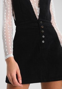 Musta korduroi nappihame, jonka päällä on läpinäkyvä valkoinen pitkähiha-toppi, jossa on kohotetut pilkut. Hame paljastaa keskijalkaan asti.