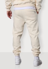 Cremefarbene Sweatpants aus Baumwollmischung mit elastischen Bündchen und einem Logo-Detail auf der Rückseite. Glatte Textur, entspannte Passform.