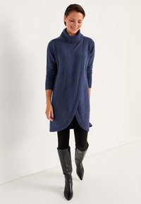 Marineblauwe, zachte gebreide coltrui met een wikkelontwerp en lange mouwen, gecombineerd met zwarte leggings en kniehoge zwarte laarzen.