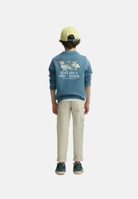 Niño con sudadera azul con gráfico y texto de dinosaurio, pantalones beige, zapatillas azules y gorra de béisbol amarilla, de espaldas sobre fondo blanco.