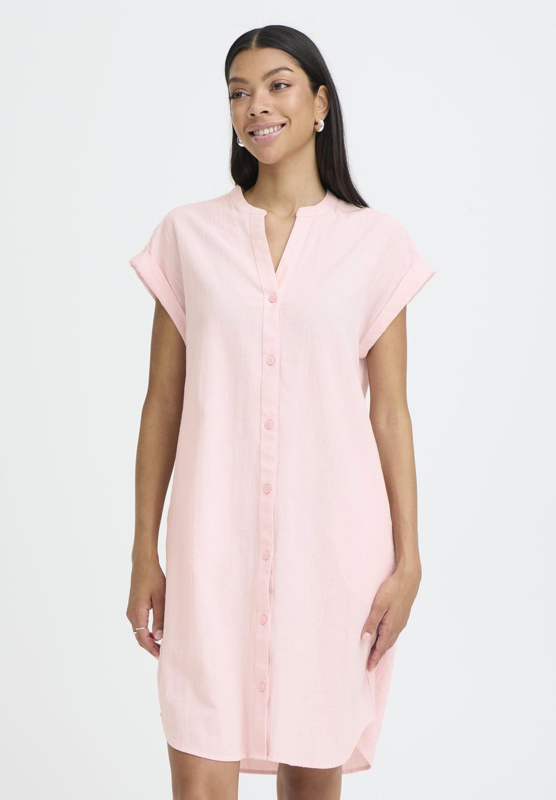 Femme aux longs cheveux foncés portant une robe rose clair à manches courtes boutonnée, souriante et regardant légèrement vers la droite.