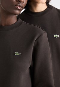 Brun sweatshirt med rund hals, prydet med en liten grønn og hvit Lacoste krokodillelogo på brystet. Mykt, teksturert stoff.