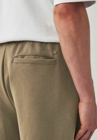Pantalons vert olive avec une texture lisse, dotés d'une poche arrière et d'un petit logo en métal. La ceinture est élastiquée pour plus de confort.