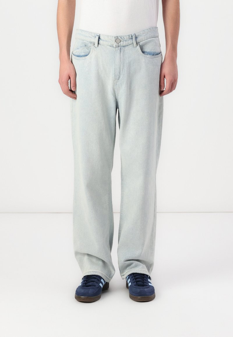 Jean en denim bleu clair, coupe ample, avec fermeture à bouton, deux poches avant et une texture lisse. Assortis à des baskets noires.