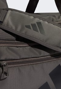 Bolsa de lona color gris oscuro con tela texturizada, que cuenta con un bolsillo con cremallera, correa ajustable y un logo negro prominente a lo largo de un lateral.