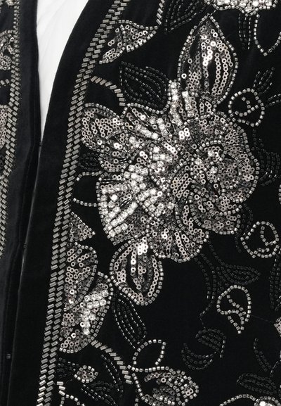 Material textil negru cu broderie florală detaliată din mărgele și paiete argintii, arătând un prim-plan al mânecii și marginii din față a unui articol vestimentar.