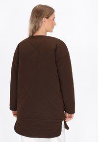 Cappotto lungo imbottito marrone con colletto rotondo, fessure laterali e una texture liscia. Il design ha una vestibilità rilassata e dettagli minimi.