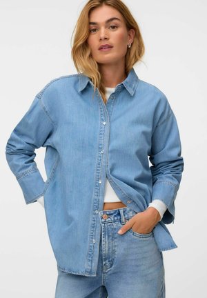 Kvinde iført en lyseblå denimskjorte over en hvid crop top og lyseblå højttaljede jeans med den ene hånd i lommen mod en ensfarvet baggrund.