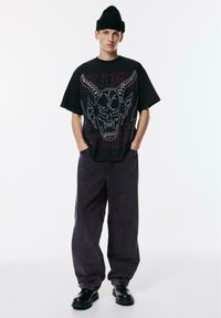 Mladic koji nosi crnu kapu, oversized crnu majicu s grafikom rogatog lubanje, široke tamne traperice i crne kožne cipele na bijeloj pozadini.