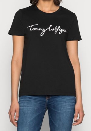 Frau trägt ein schwarzes Kurzarm-T-Shirt mit dem in weißer Schrift geschriebenen Text ""Tommy Hilfiger"" und blaue Jeans.