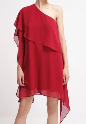 Kobieta nosząca czerwoną, asymetryczną sukienkę bez rękawów, z jednym ramieniem zakrytym i warstwową przezroczystą tkaniną opadającą z jednej strony.