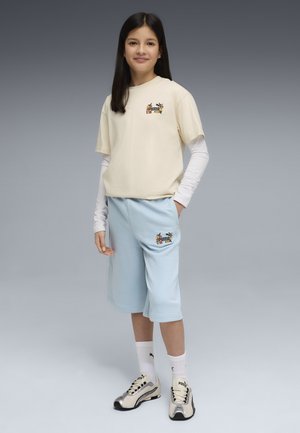 Młoda dziewczyna stoi z jedną ręką w kieszeni, ubrana w beżową koszulę z krótkim rękawem na białym długim rękawie i jasnoniebieskie szorty z trampkami.