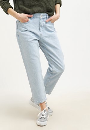 Osoba nosząca jasnoniebieskie, wysokie dżinsy z rękami w kieszeniach, w połączeniu z srebrnymi, metalicznymi trampkami i ciemnozielonym dzianinowym swetrem.