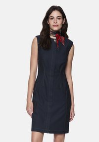 Marineblaues ärmelloses Kleid aus Denim mit vertikalen Nähten, zwei vorderen Reißverschlusstaschen und kontrastierenden goldenen Steppnähten. Roter, gemusterter Schal als Akzent.