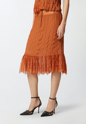 Jupe midi plissée en orange brûlé avec une bordure en dentelle à l'ourlet. Portée avec des sandales à talons noires et un top ajusté en texture.