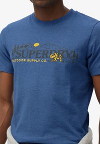 Blå t-shirt med teksten "SUPERDRY OUTDOOR SUPPLY CO.", gul sol, bjørn, bjerg og træ grafik på brystet.