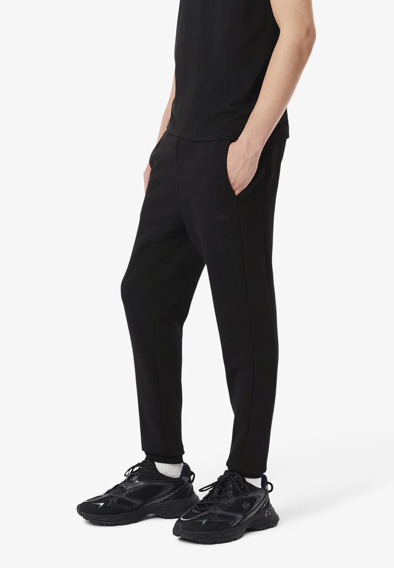Pantalons de survêtement noirs en tissu doux, dotés de poches latérales, d'une coupe slim et d'un détail de logo discret. Associés à des chaussures de sport noires.