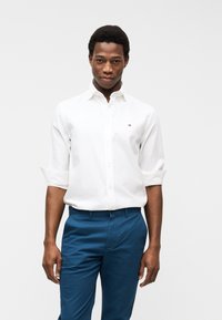 Νεαρός άνδρας που φοράει λευκό πουκάμισο με κουμπιά και μανίκια γυρισμένα, και μπλε παντελόνι, στέκεται μπροστά σε ένα λευκό, απλό φόντο.