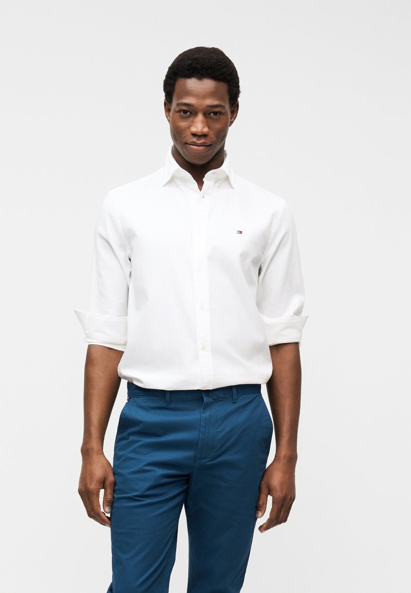 Νεαρός άνδρας που φοράει λευκό πουκάμισο με κουμπιά και μανίκια γυρισμένα, και μπλε παντελόνι, στέκεται μπροστά σε ένα λευκό, απλό φόντο.