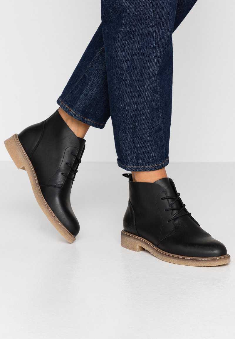 Bottes montantes en cuir noir avec un bout arrondi, un design à lacets et une semelle texturée de couleur claire. Portées avec un pantalon en denim foncé.